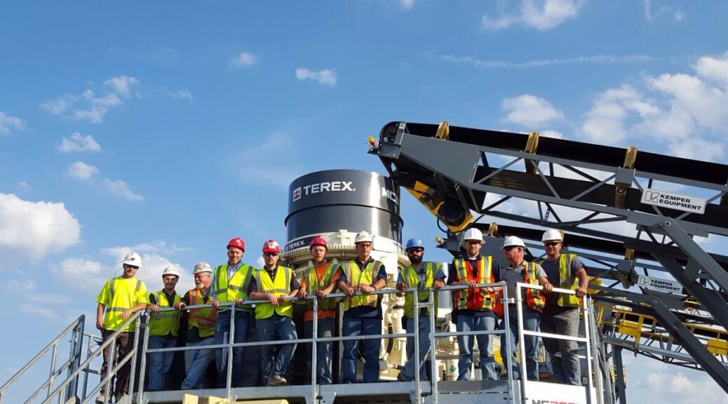 A group of people stands on an equipment platform wearing safety gear, hard hats, and reflective vests.